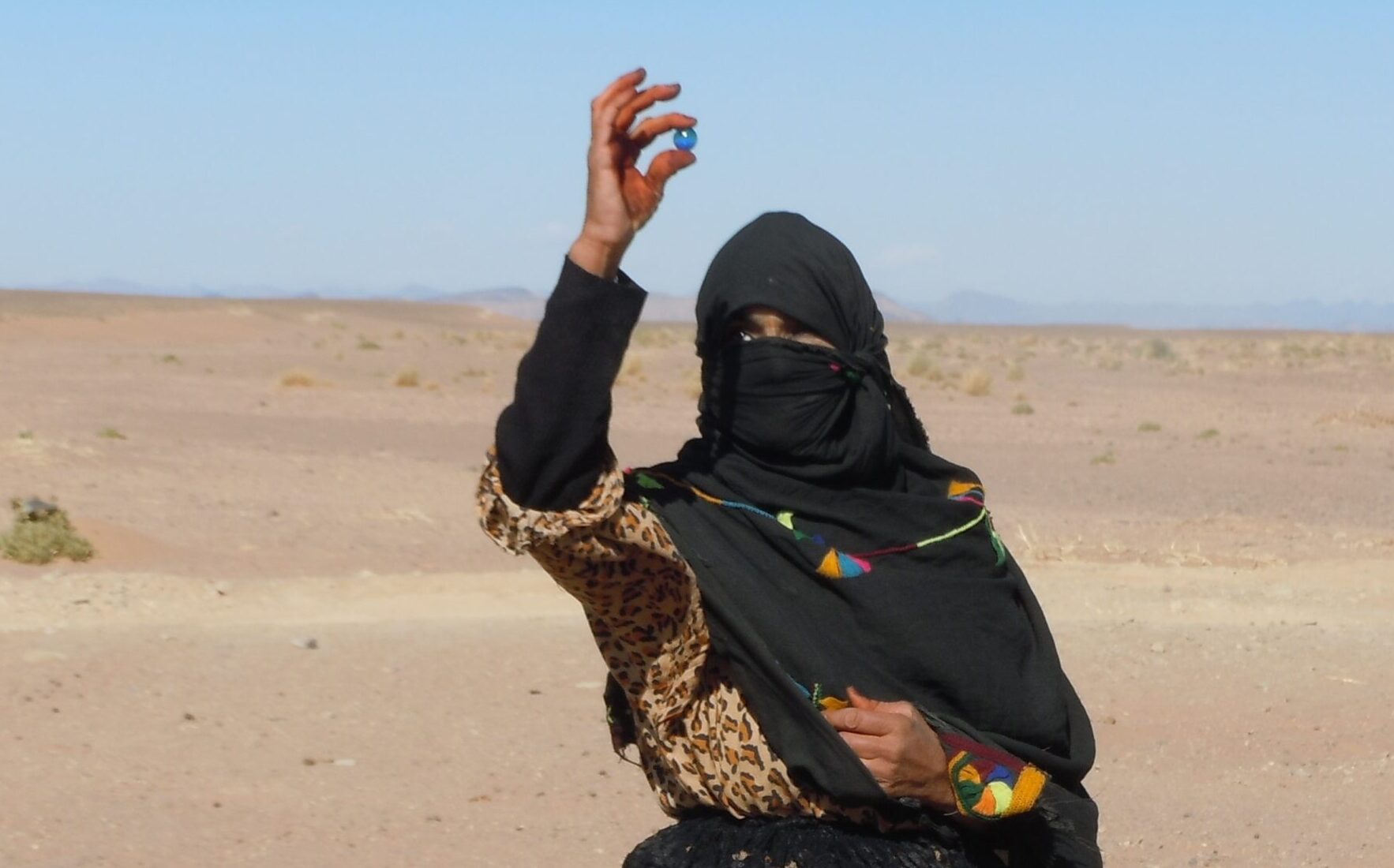 A women wearing a black hijab stands in the arid desert landscape of the Sahara. She is holding a small blue marble in her right hand.